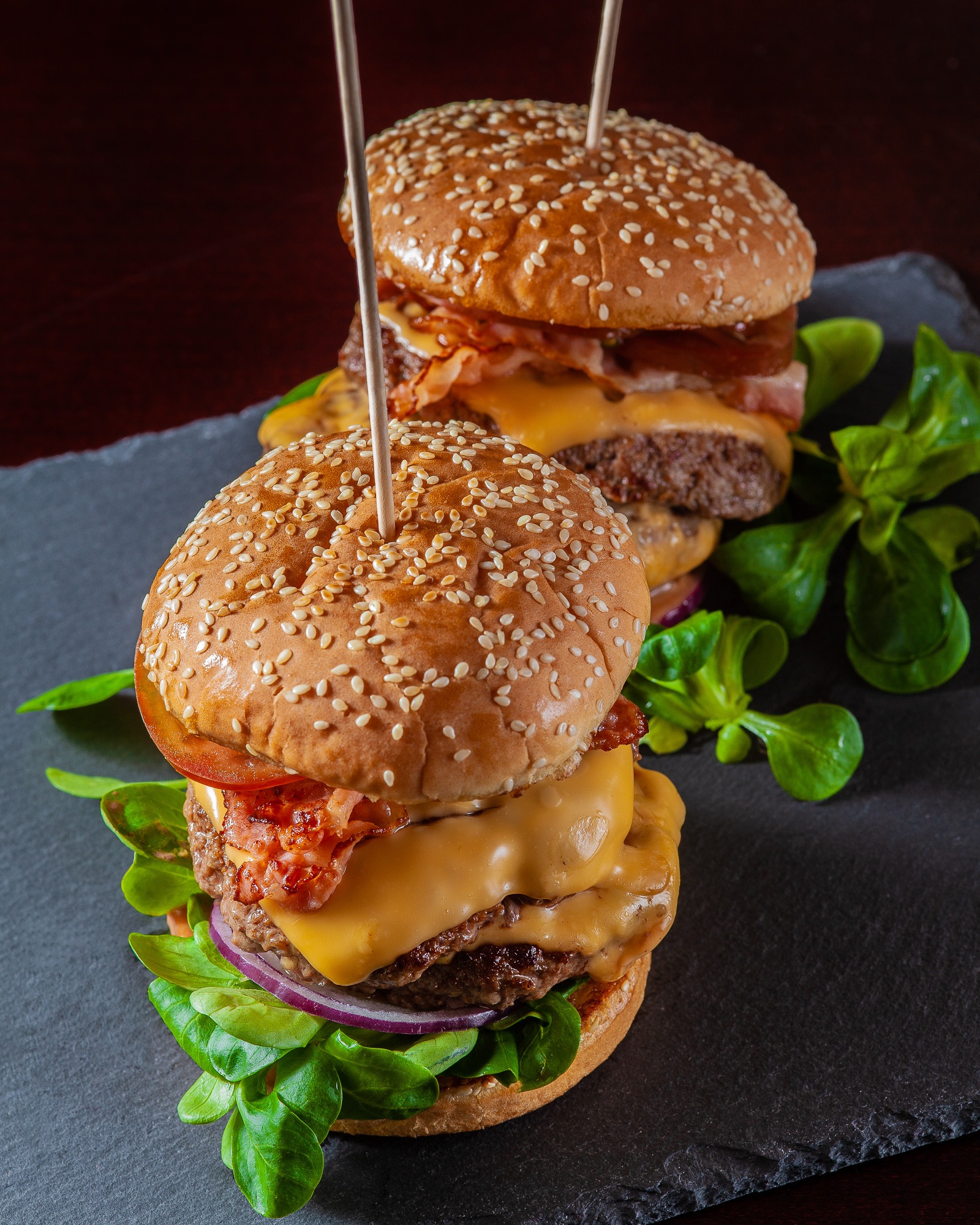 Dwa burgery z wołowiną, serem cheddar, bekonem, pomidorem, czerwoną cebulą i sałatą, spięte wykałaczkami, na czarnym kamiennym talerzu.