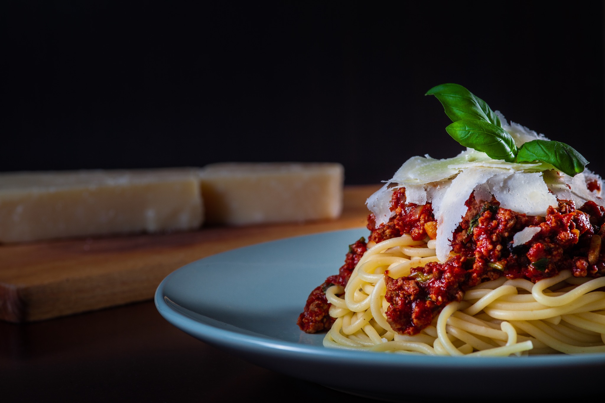 Spaghetti bolognese na niebieskim talerzu, posypane parmezanem i udekorowane listkami bazylii, z kawałkami sera w tle.