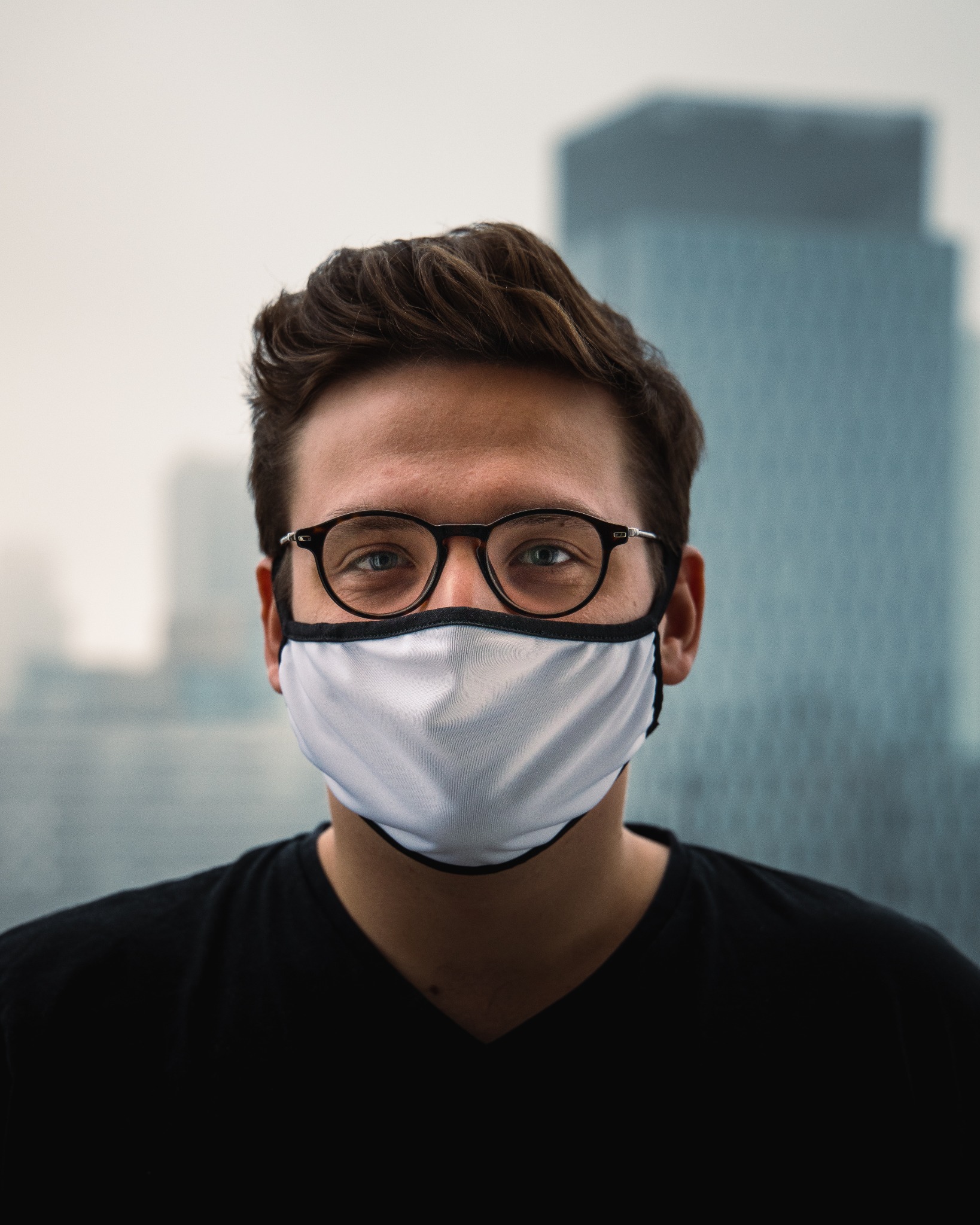 Portret mężczyzny w okularach i białej maseczce ochronnej na tle miejskiego krajobrazu z wysokimi budynkami.