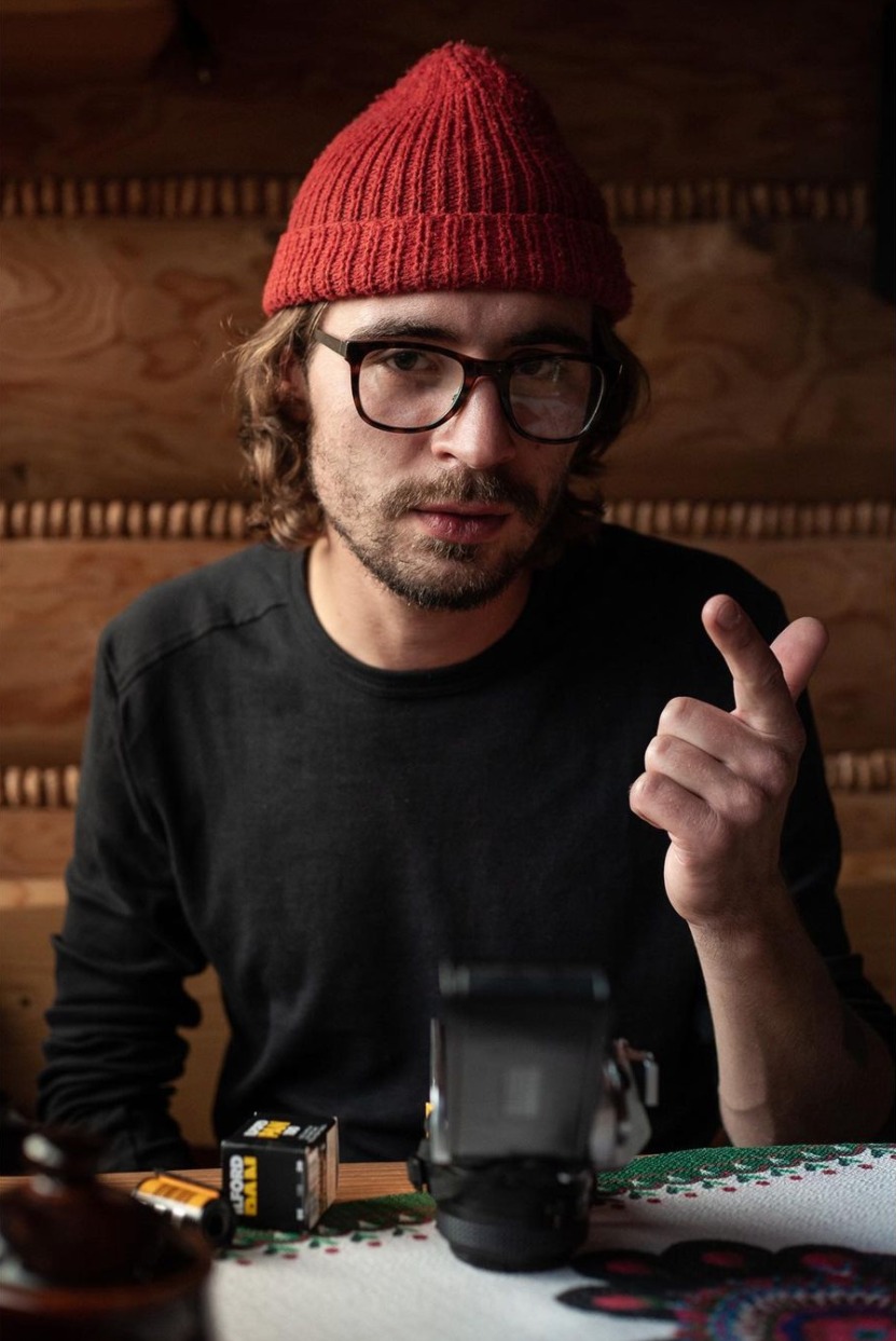 Portret fotografa w czerwonej czapce i okularach, z aparatem analogowym i filmami na stole z haftowanym obrusem, na tle drewnianej ściany.