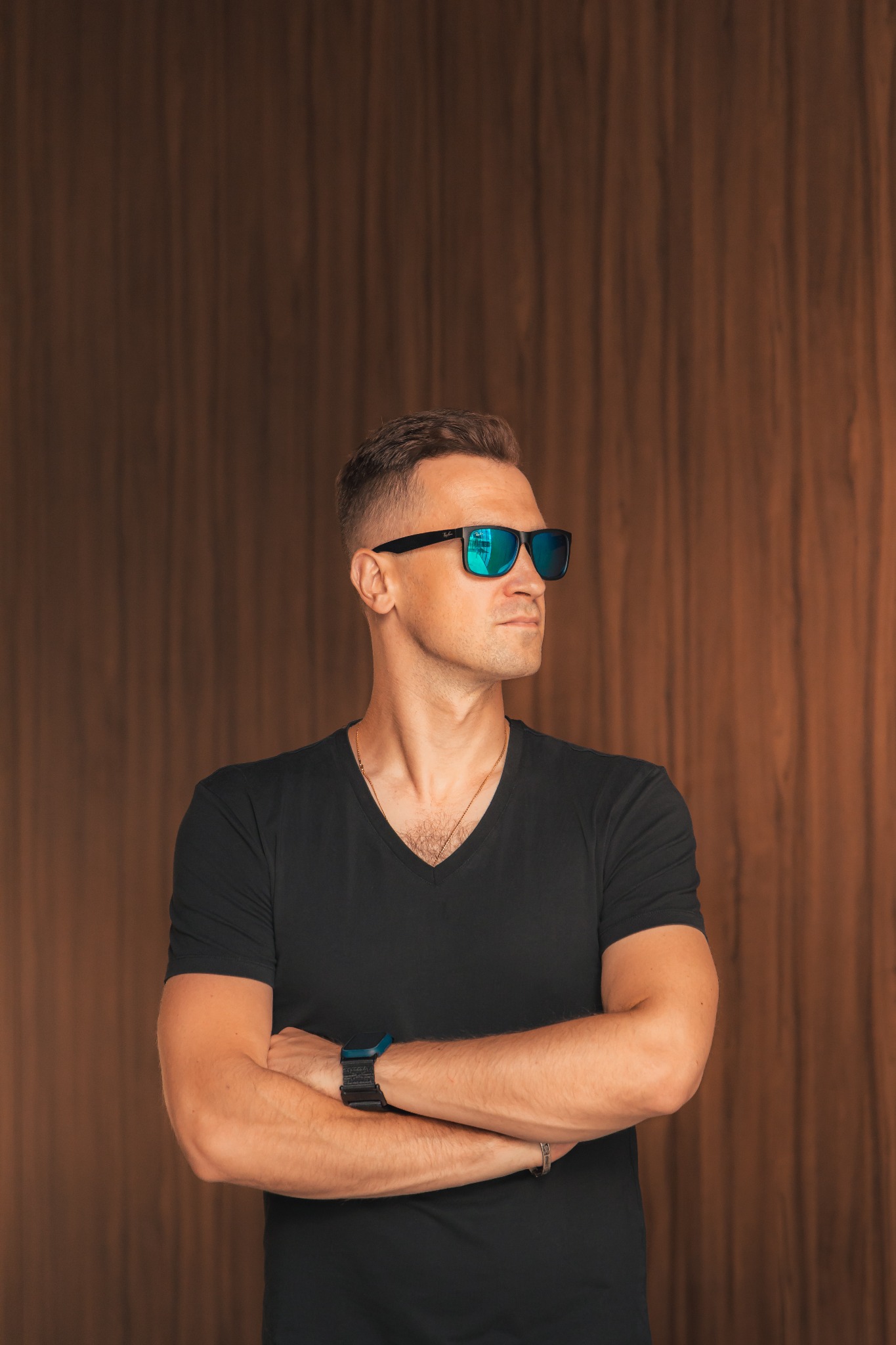 Mężczyzna w czarnej koszulce z założonymi rękami, noszący okulary przeciwsłoneczne z niebieskim odbiciem, stojący na tle drewnianej ściany.