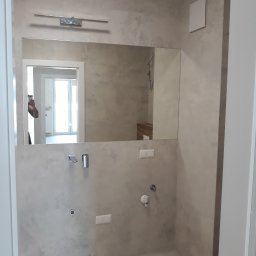 OLVIT - Wykończona łazienka w odcieniach beżu z dużym lustrem nad umywalką, widoczne baterie, oświetlenie nad lustrem i wentylator, w odbiciu lustra widać fragment innego pomieszczenia.