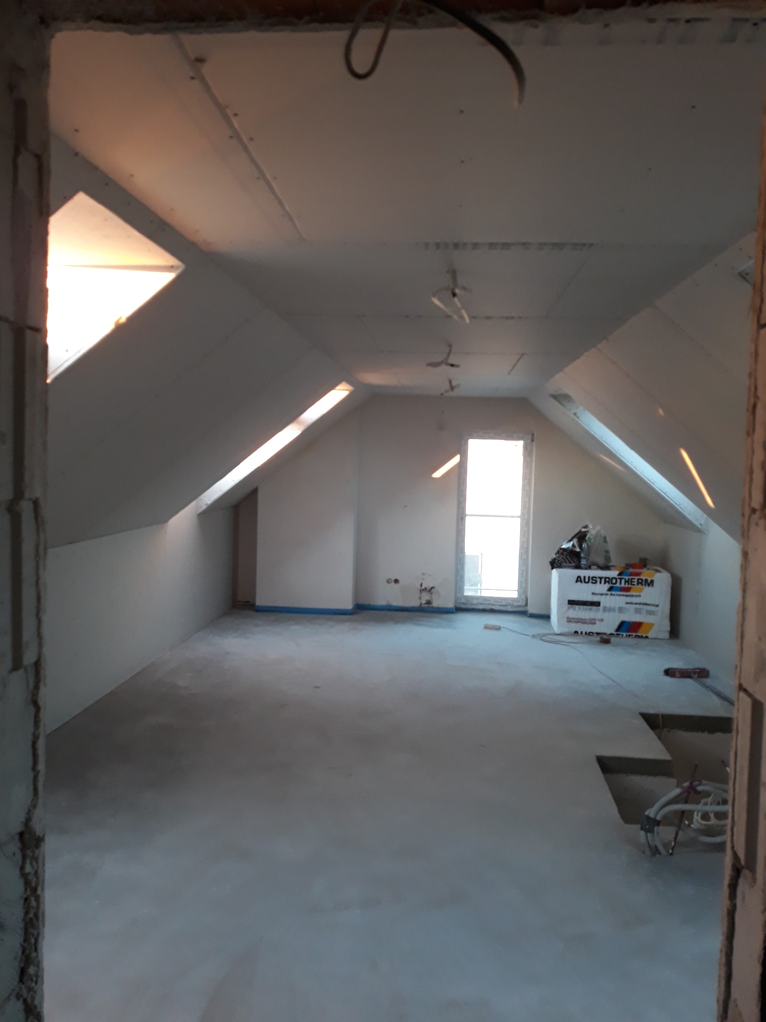 Poddasze w trakcie wykańczania, widoczne okna dachowe, ściany z płyt gipsowo-kartonowych, izolacja termiczna Austrotherm i przewody elektryczne wystające z podłogi.