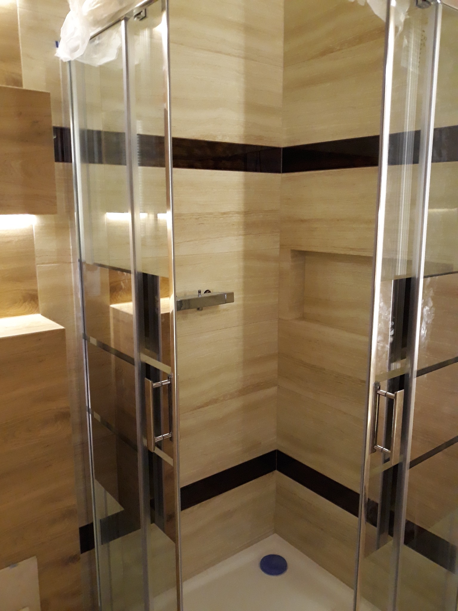 Kabina prysznicowa z przesuwnymi drzwiami, wykończona płytkami w odcieniach beżu z ciemnobrązowymi akcentami, widoczna półka w ścianie i niebieski odpływ w brodziku.