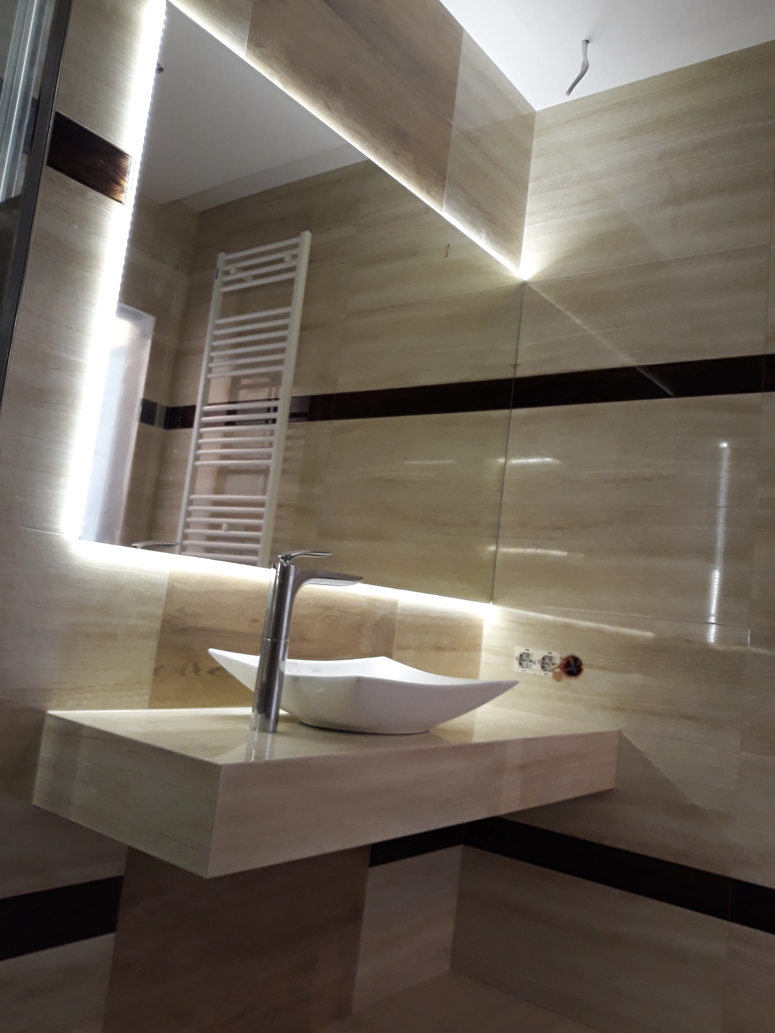 Biała, asymetryczna umywalka nablatowa z chromowaną baterią na blacie z imitacji marmuru w łazience z beżowymi płytkami i podświetlanym lustrem.