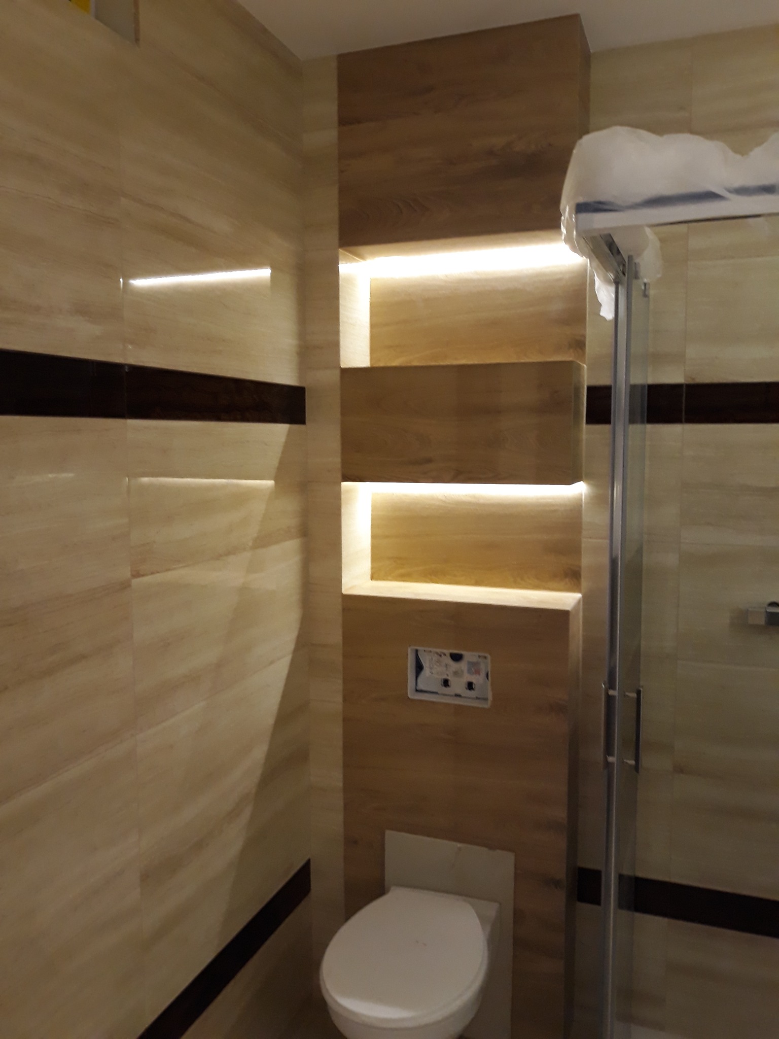 Wykończona łazienka z beżowymi płytkami imitującymi drewno, zabudowa z półkami podświetlonymi ledami, wisząca miska WC i częściowo widoczna kabina prysznicowa.