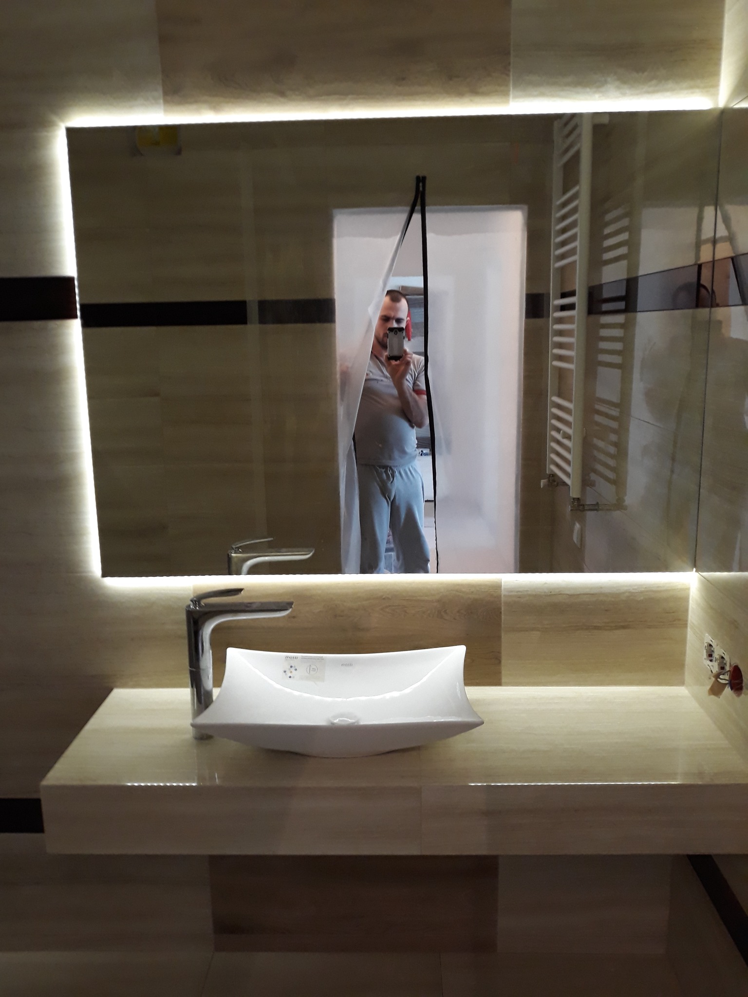 Nowoczesna łazienka z podświetlanym lustrem, umywalką w kształcie łodzi i grzejnikiem drabinkowym, widoczna osoba robiąca zdjęcie w lustrze.