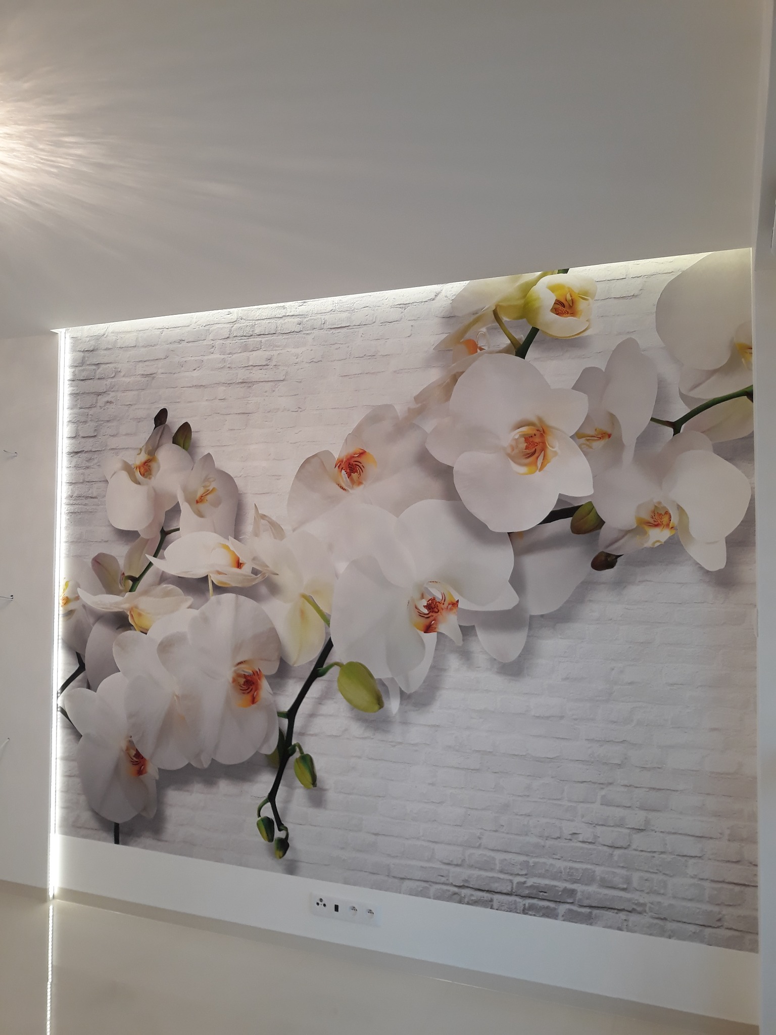 Fototapeta z motywem białych orchidei na ścianie imitującej białą cegłę, oświetlona listwą LED, widok z dołu.