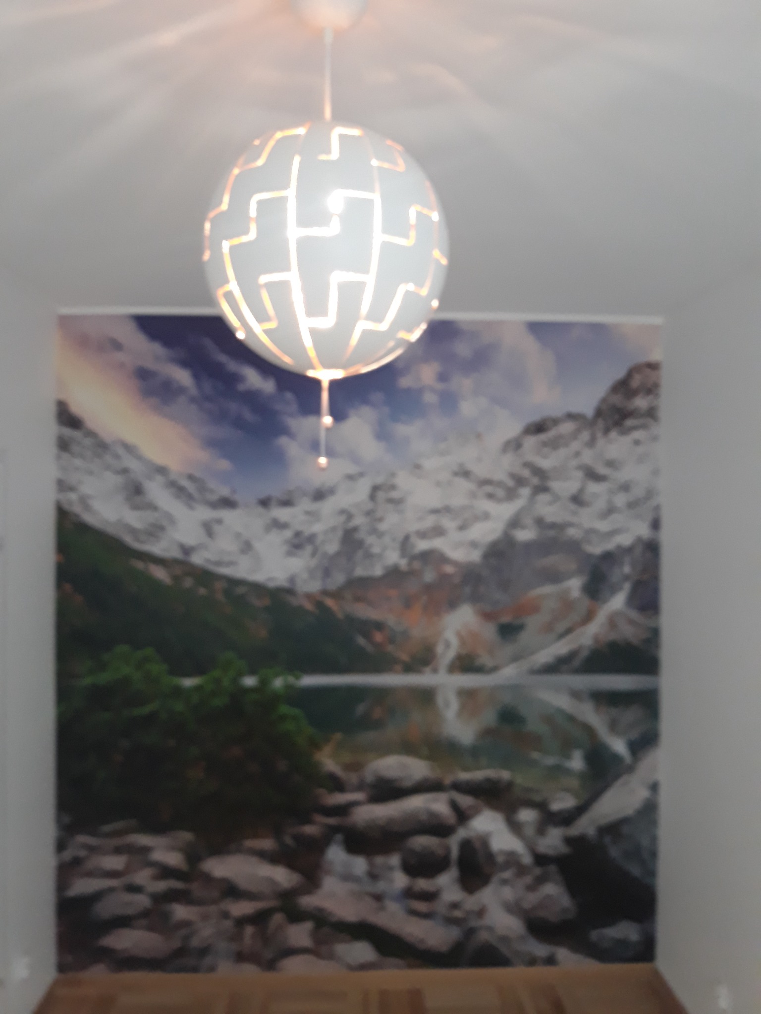 Wnętrze pokoju z dekoracyjną lampą sufitową w kształcie kuli i fototapetą z widokiem górskiego jeziora.