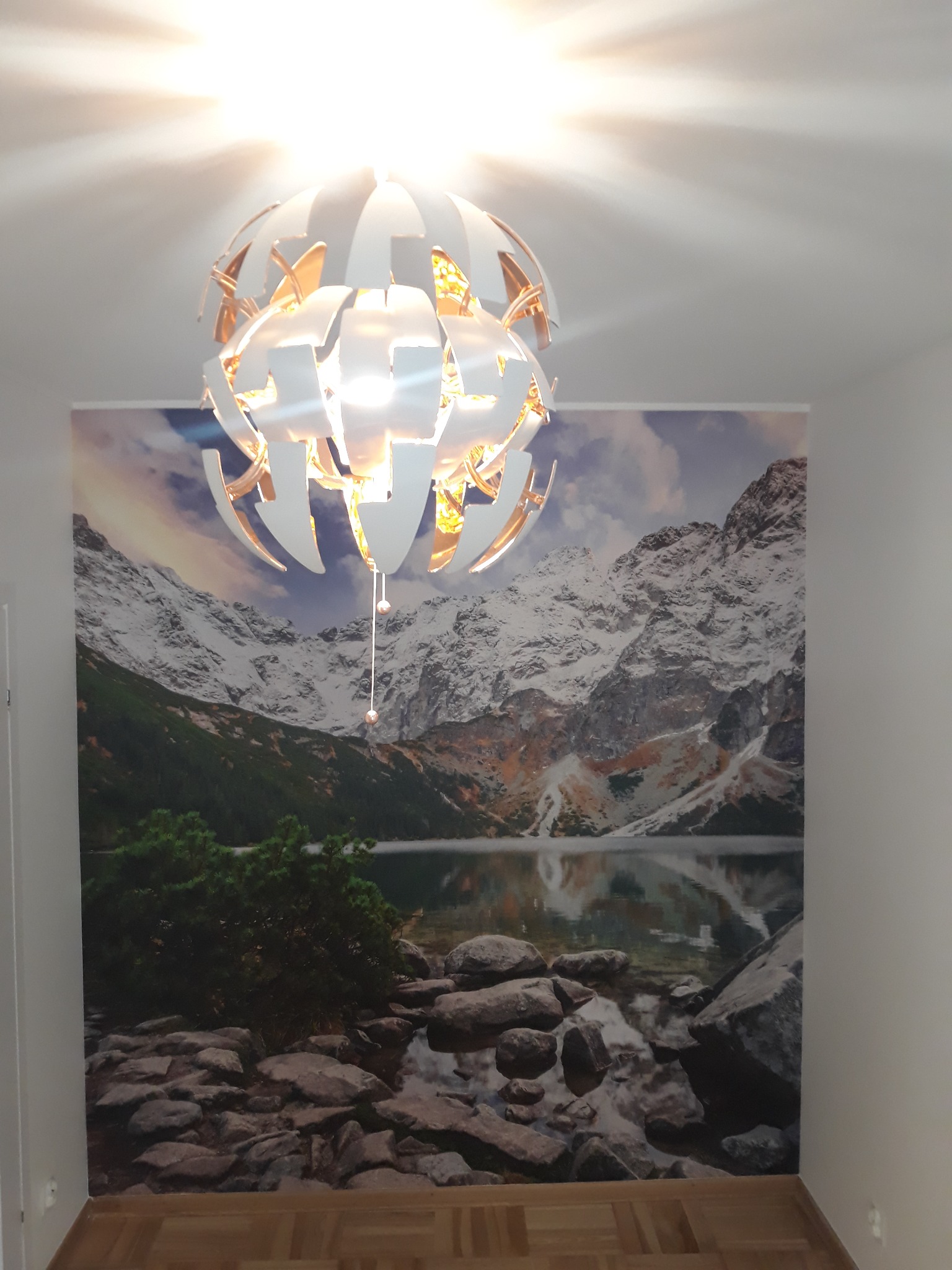 Nowoczesna lampa sufitowa w kształcie kuli z białymi i złotymi elementami, zawieszona w pokoju z fototapetą przedstawiającą górski krajobraz z jeziorem i kamienistym brzegiem.