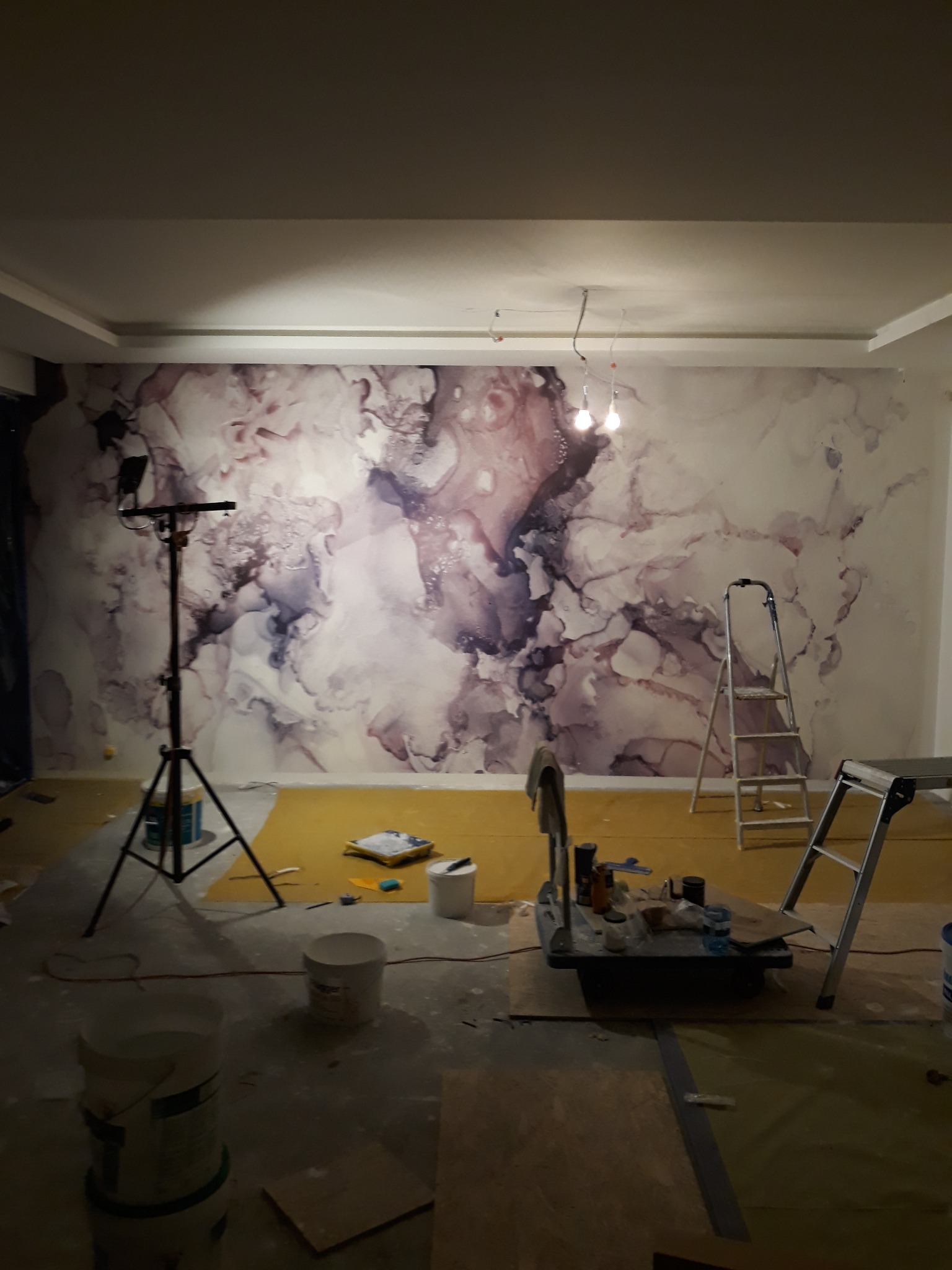Ściana po malowaniu z efektem marmuru, widoczne narzędzia malarskie, drabina, lampa oświetleniowa na statywie i wiaderka z farbą na podłodze zabezpieczonej folią.