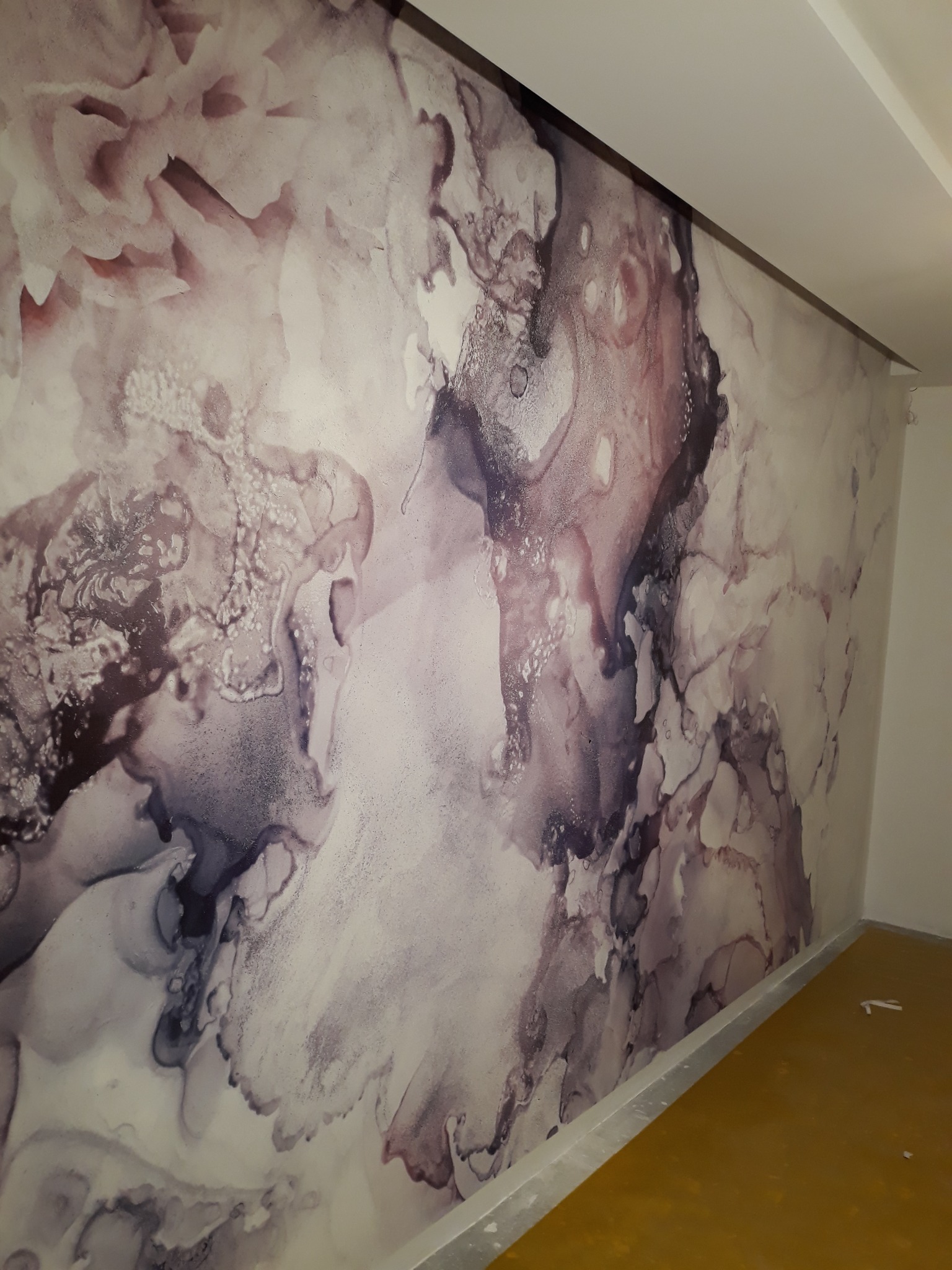 Ściana pokryta tapetą z abstrakcyjnym wzorem przypominającym marmur w odcieniach szarości, bieli i fioletu, widoczny fragment podłogi w kolorze musztardowym.