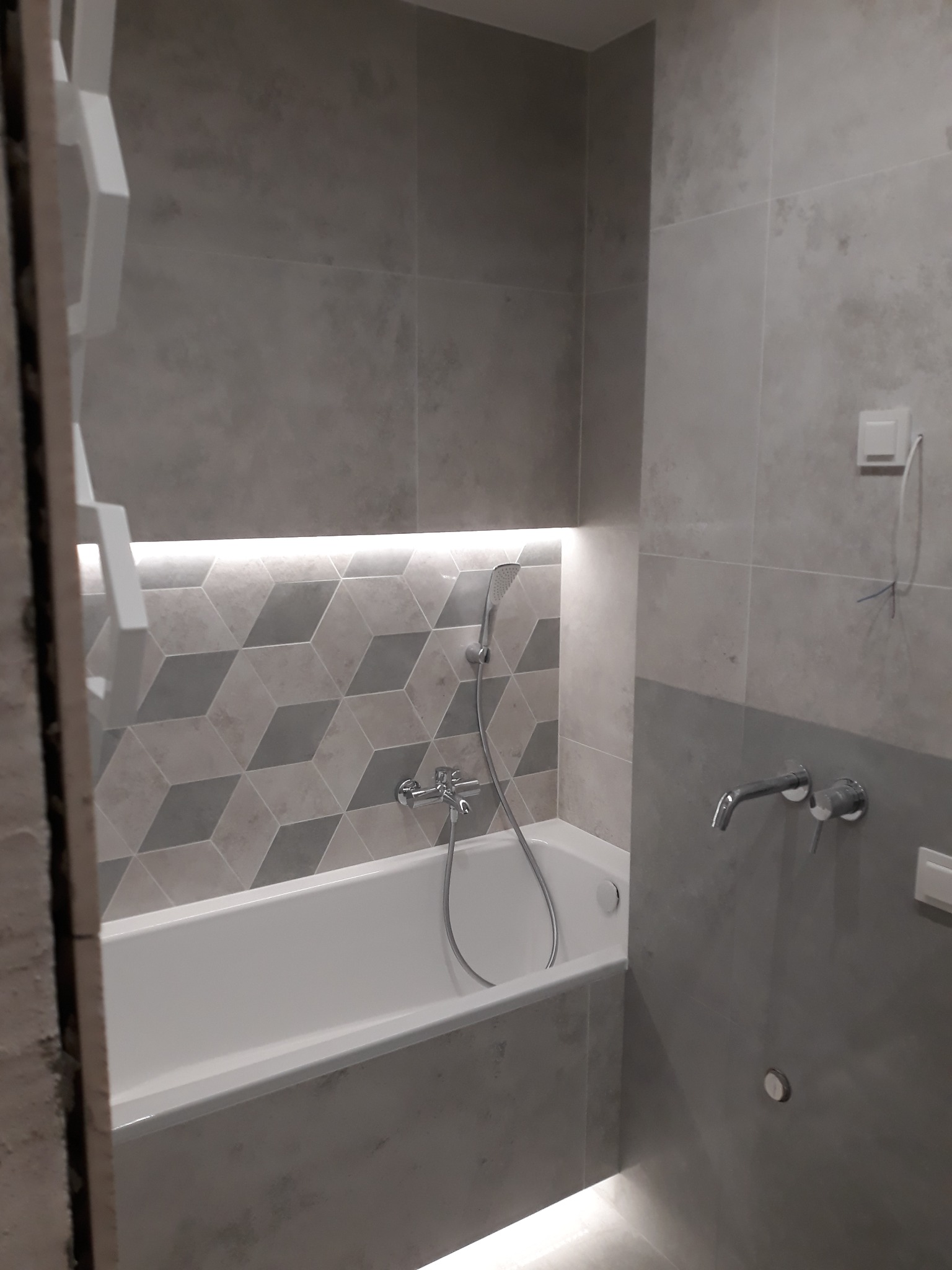 Wykończona łazienka z szarą płytką imitującą beton, białą wanną, baterią podtynkową i podświetleniem LED nad geometryczną mozaiką za wanną.