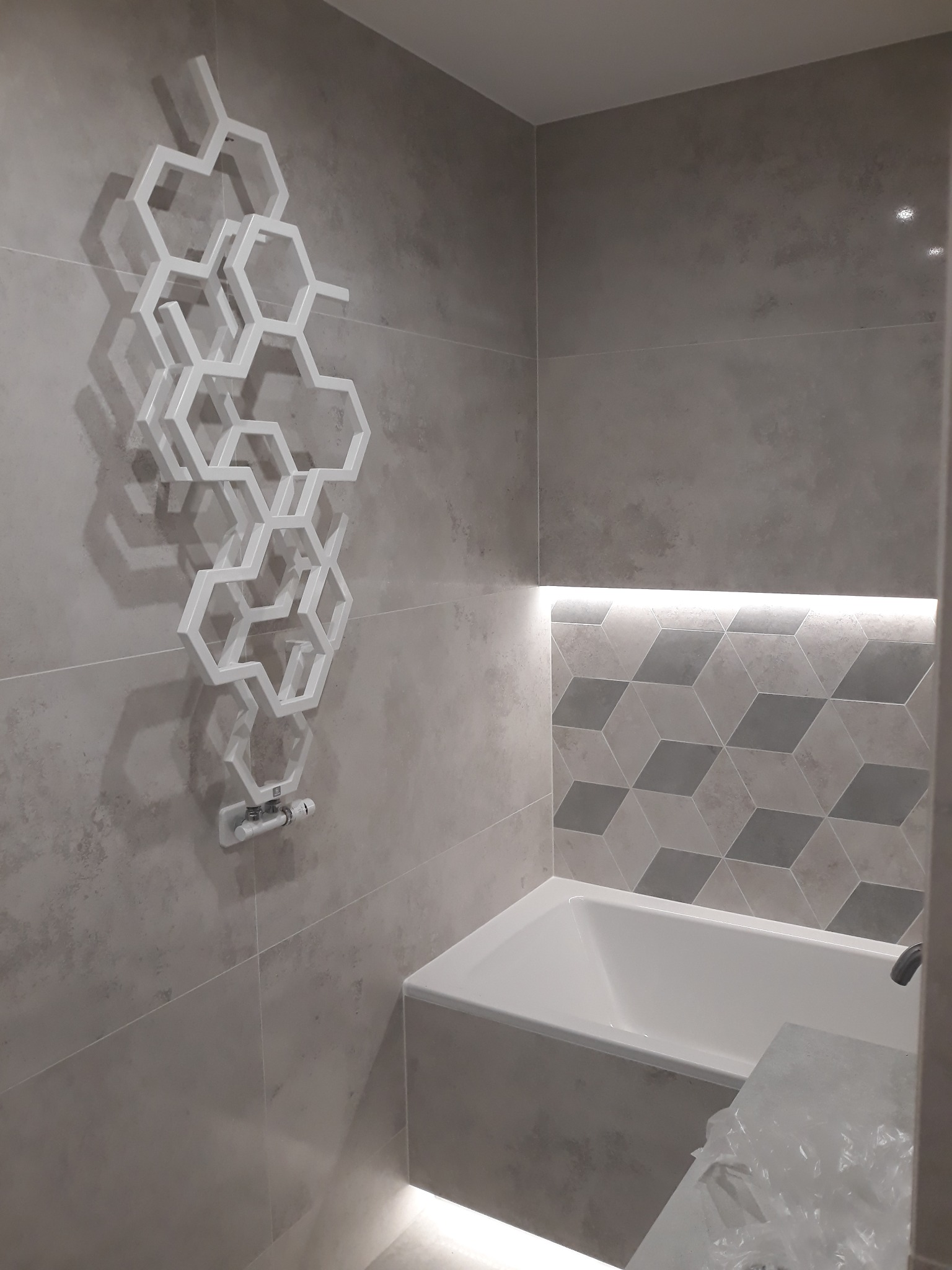 Nowoczesna łazienka w szarościach z designerskim, białym grzejnikiem drabinkowym o heksagonalnej strukturze, podświetlaną wnęką nad wanną i geometrycznym wzorem płytek.