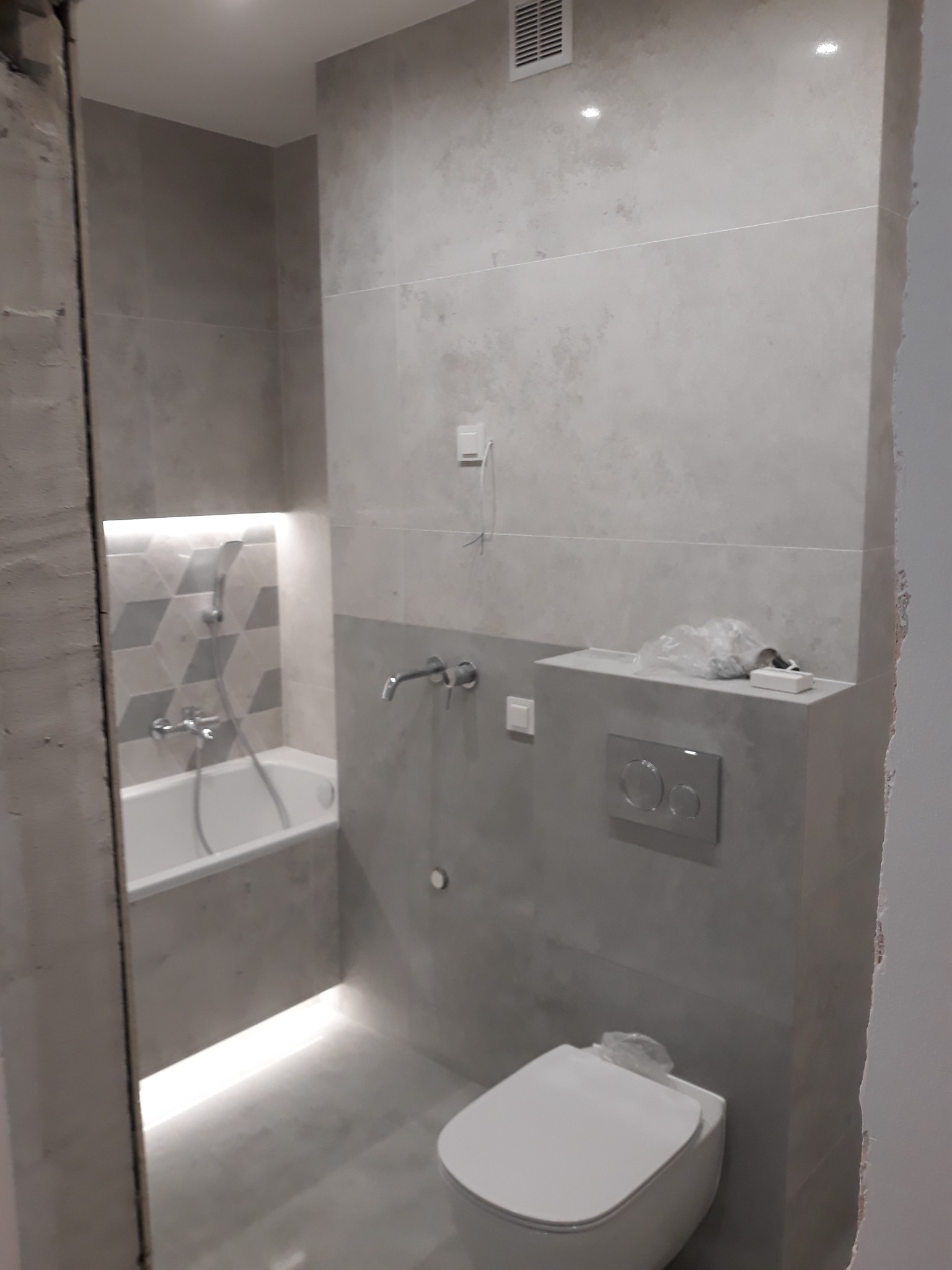 Nowoczesna łazienka w trakcie wykańczania: szare płytki imitujące kamień, podświetlana wnęka z wanną i geometrycznym wzorem, wisząca toaleta oraz fragment niezabezpieczonej ściany w stanie surowym.