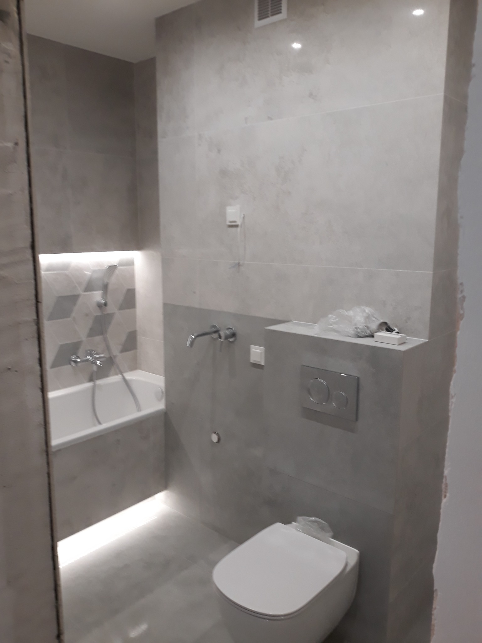 Nowoczesna łazienka w odcieniach szarości z podświetlaną wanną, wiszącą toaletą i geometrycznymi płytkami na ścianie wnęki prysznicowej.