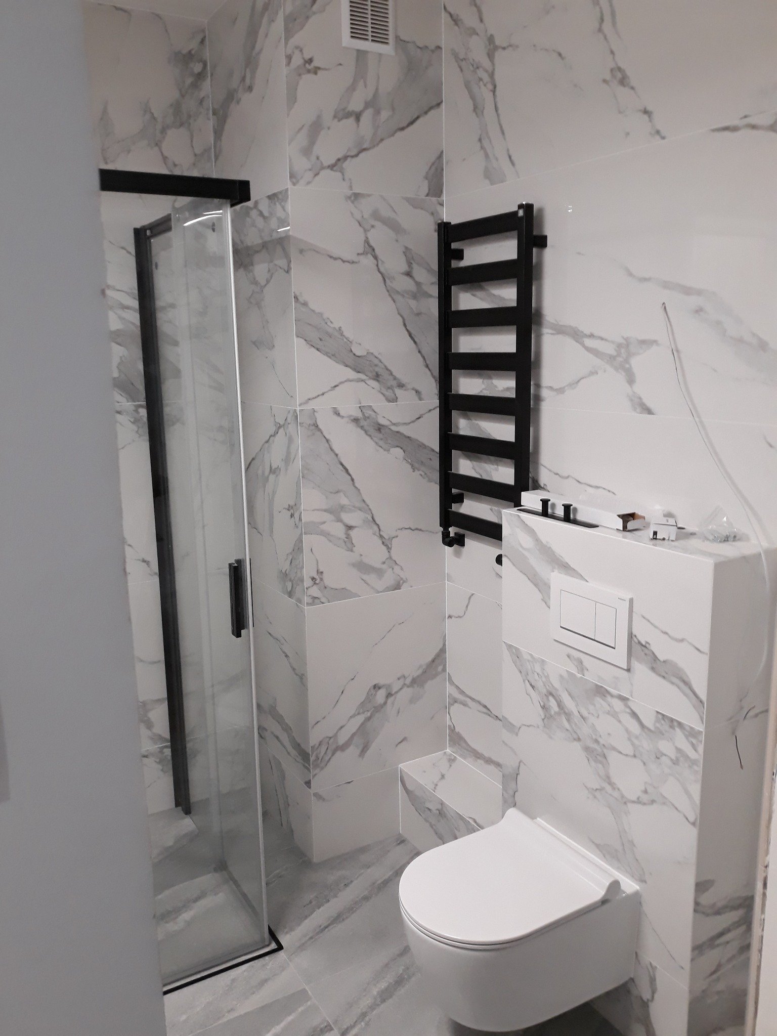 Nowoczesna łazienka z kabiną prysznicową z czarnymi ramami, wiszącą toaletą, grzejnikiem drabinkowym w czarnym kolorze i marmurowymi płytkami na ścianach i podłodze.