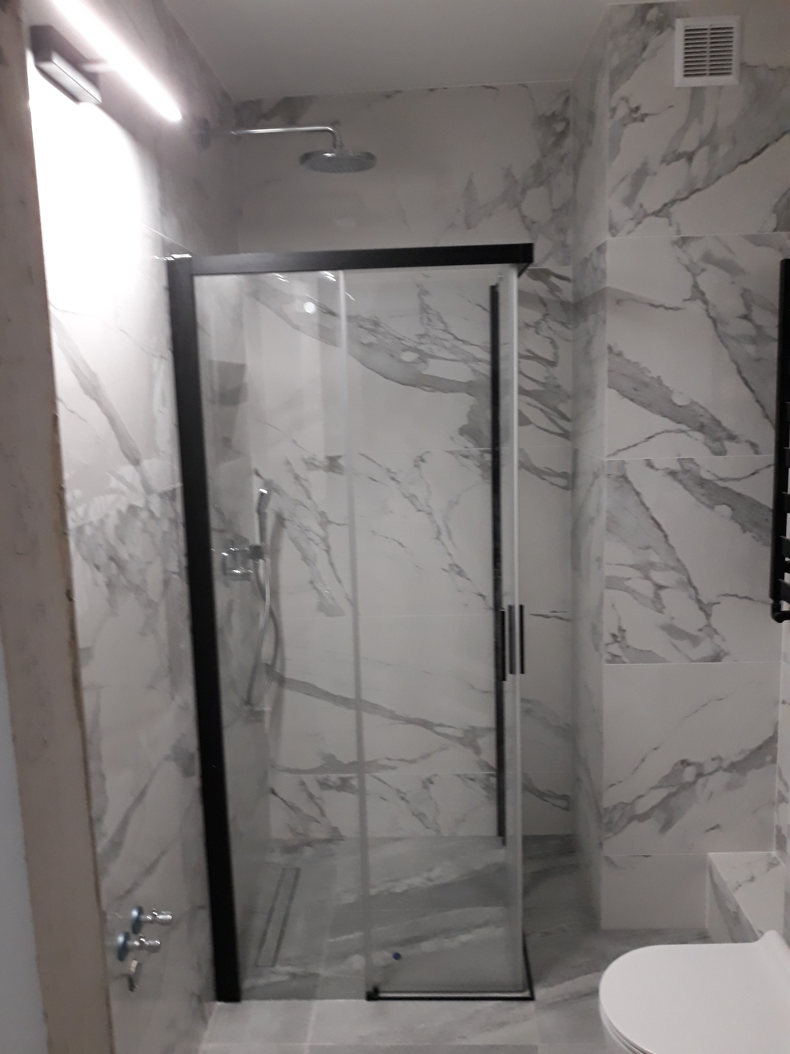 Nowoczesna kabina prysznicowa z czarną ramą i marmurkowymi płytkami w łazience w Warszawie, widoczny fragment toalety i grzejnika.