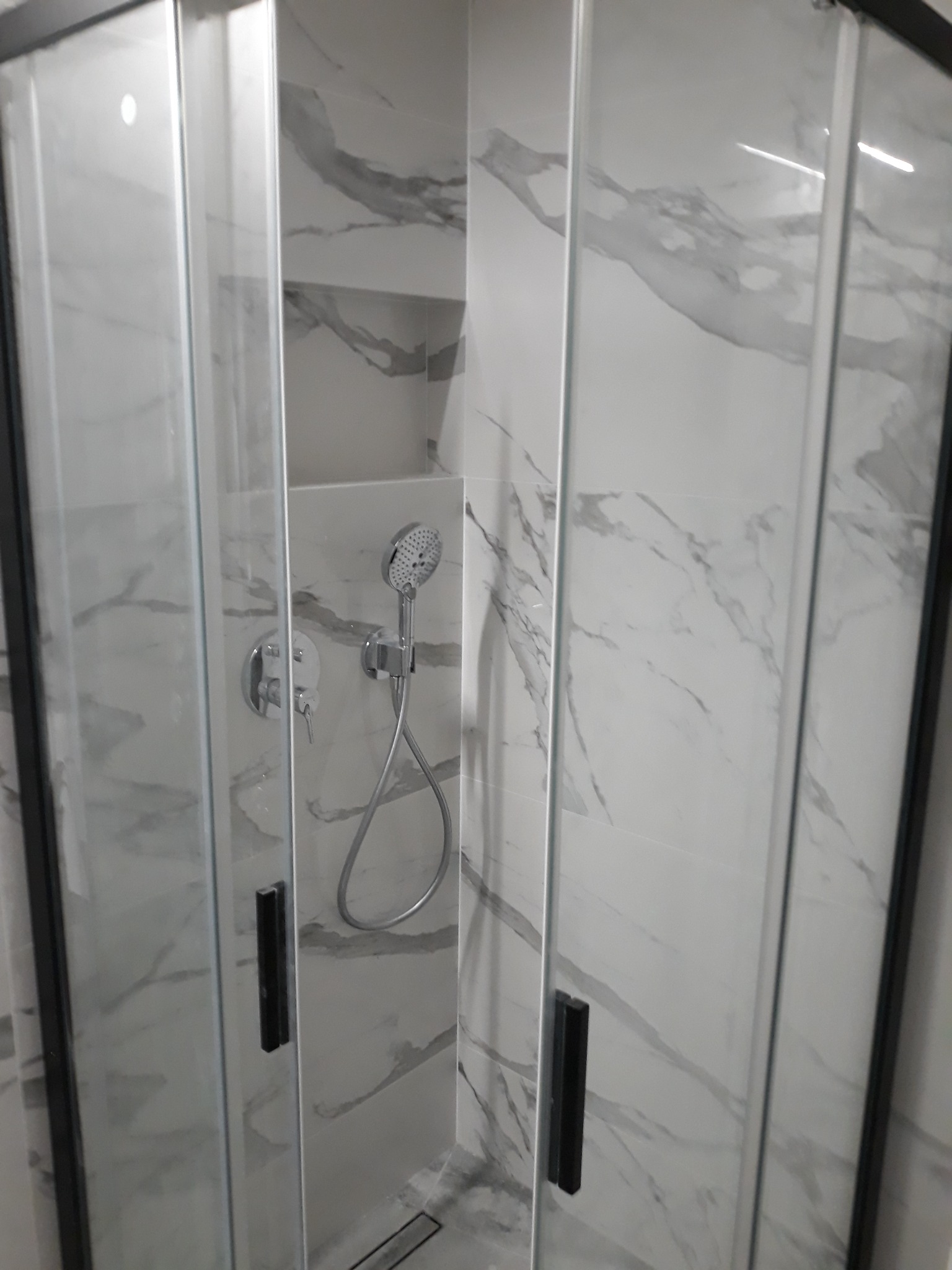 Kabina prysznicowa z czarnymi ramami i szklanymi drzwiami na tle płytek imitujących marmur, widoczna bateria i słuchawka prysznicowa.