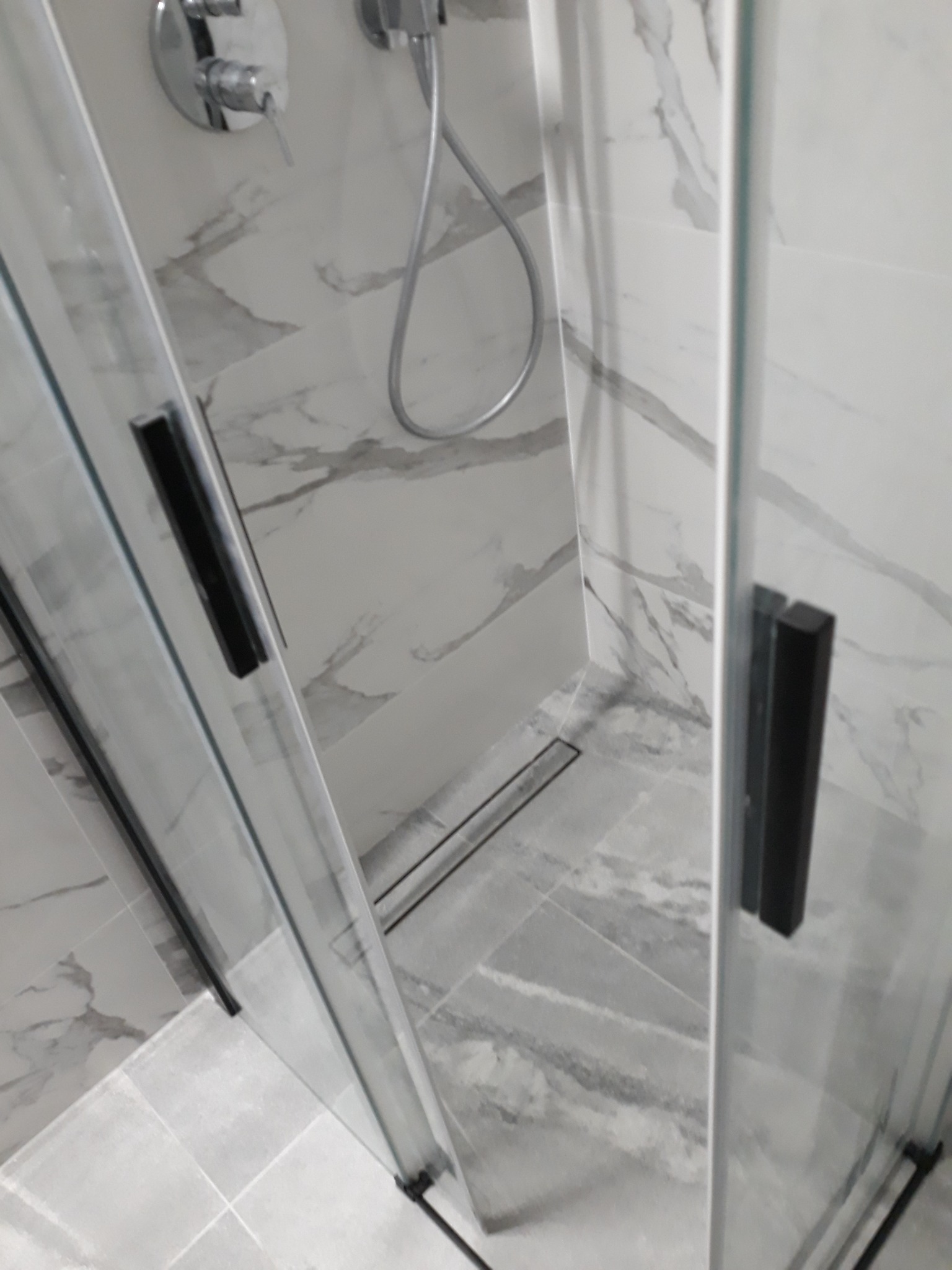 Szklana kabina prysznicowa z czarnymi uchwytami i odpływem liniowym, wykończona płytkami imitującymi marmur w odcieniach szarości.