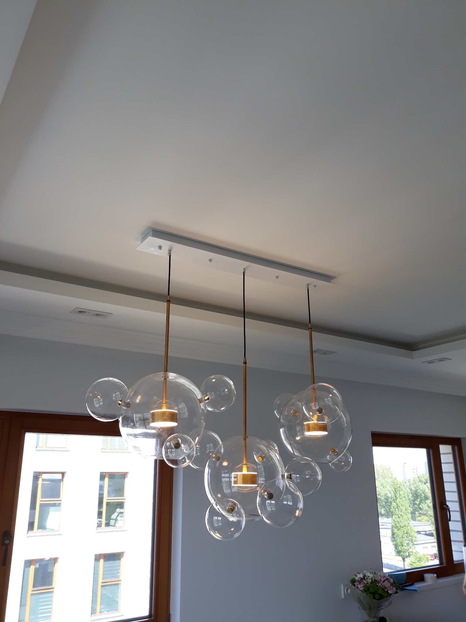 Nowoczesny, designerski żyrandol z przezroczystych, szklanych kul zawieszony na złotych prętach, oświetlający wnętrze z widokiem na budynek mieszkalny przez drewniane ramy okienne.