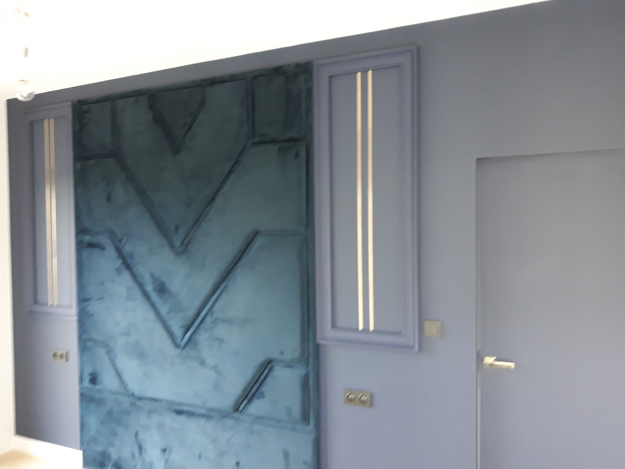 Niebieska ściana z geometrycznym wzorem i dekoracyjnymi pionowymi panelami z jasnymi paskami, obok szarych drzwi z klamką