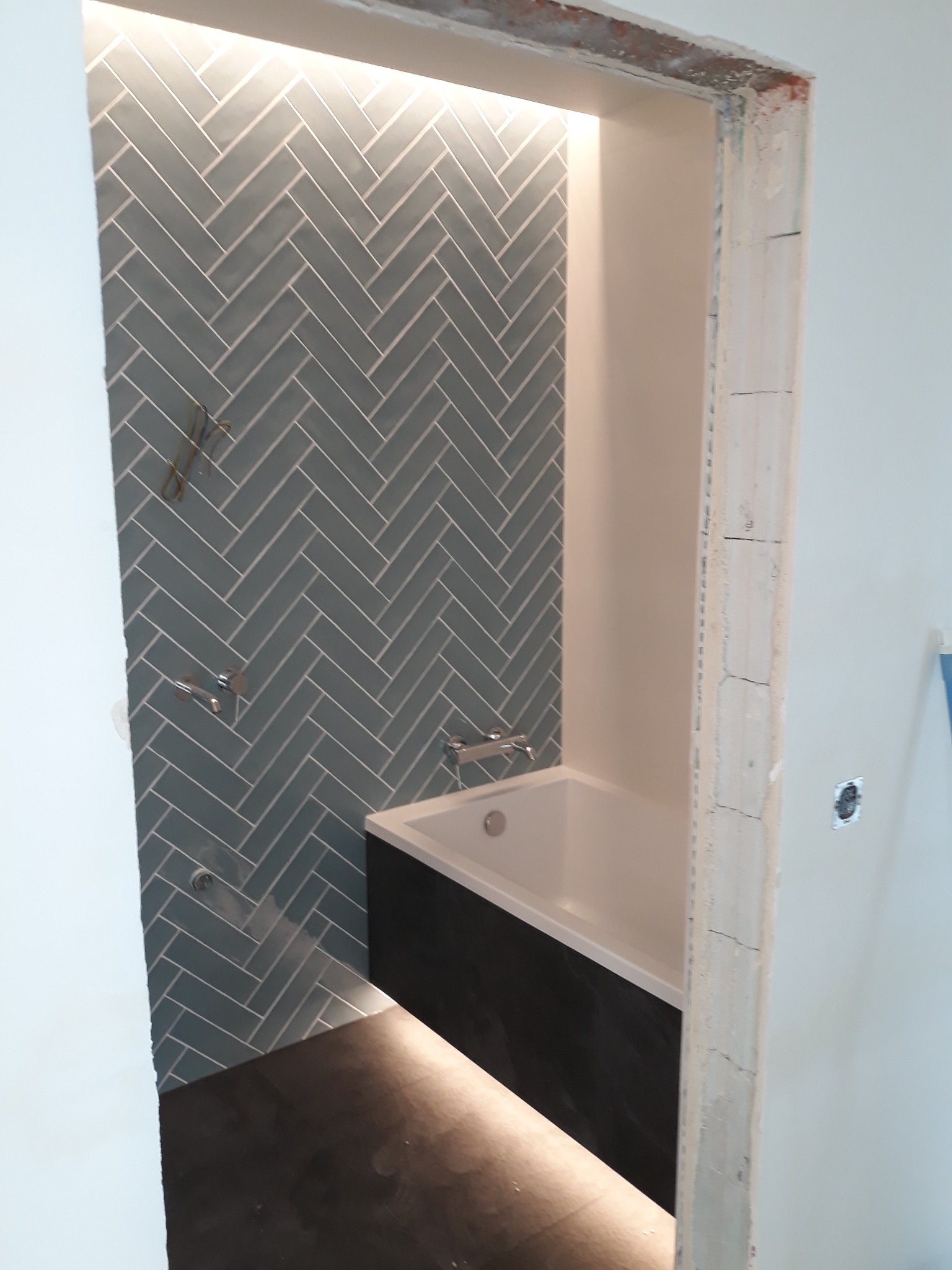 Wnętrze łazienki w trakcie prac wykończeniowych: ściana wyłożona niebiesko-szarą glazurą ułożoną w jodełkę, widoczna wanna z czarną obudową i surowa ościeżnica.