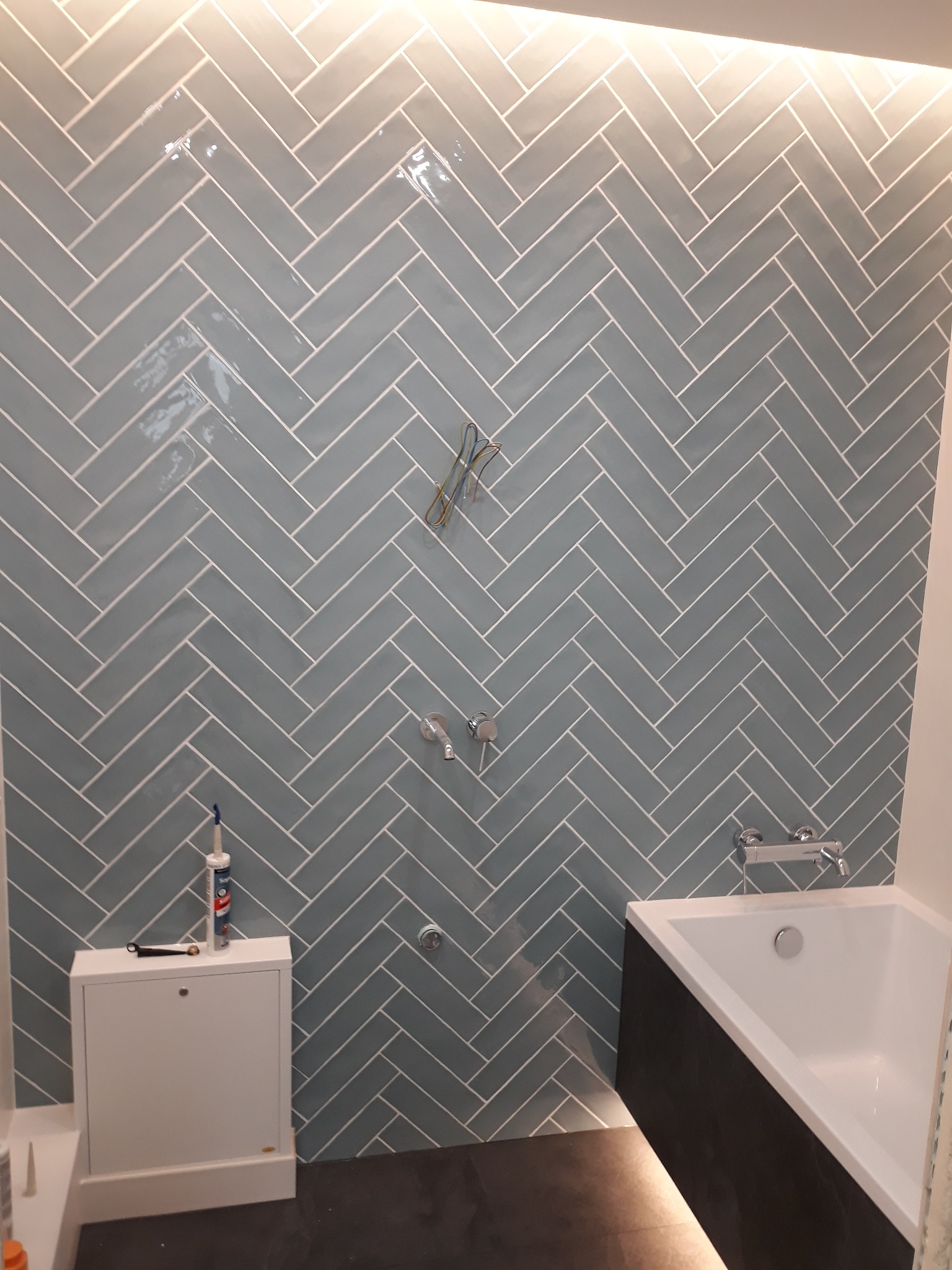 Ściana w łazience wyłożona błyszczącymi, błękitnymi płytkami ułożonymi w jodełkę, widoczna bateria wannowa, syfon, biała szafka z silikonem i narzędziem na górze, oraz fragment wanny.