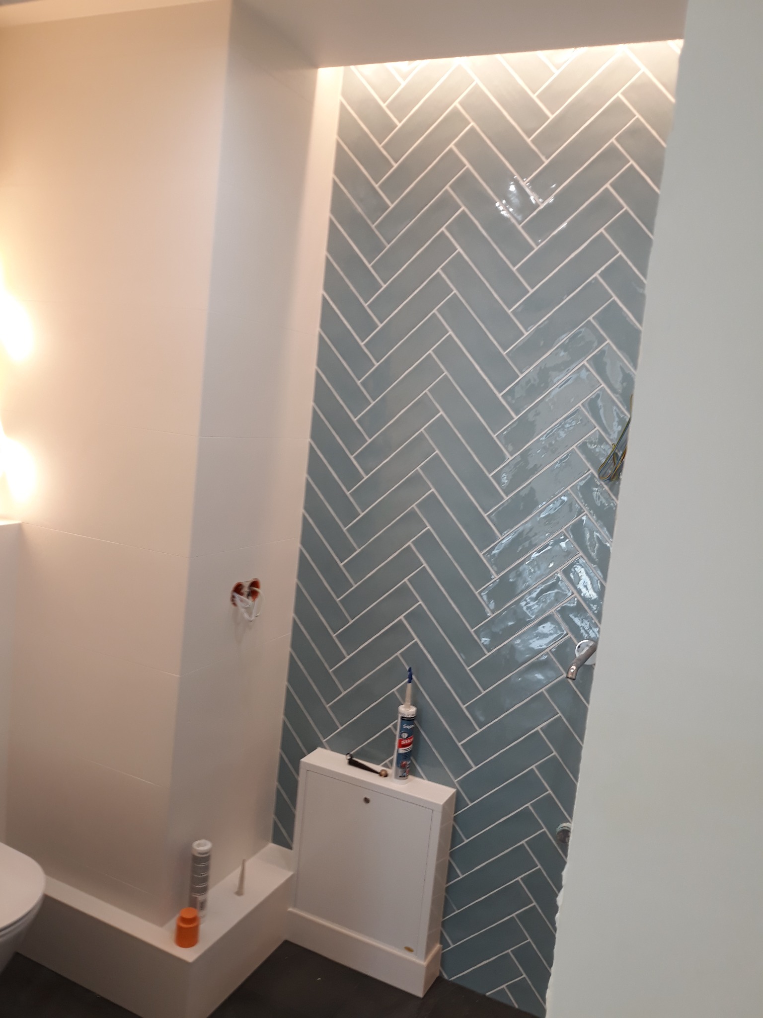 Fragment łazienki w trakcie wykańczania, widoczne ułożone w jodełkę błękitne płytki, biała zabudowa z dostępem do instalacji oraz akcesoria montażowe.