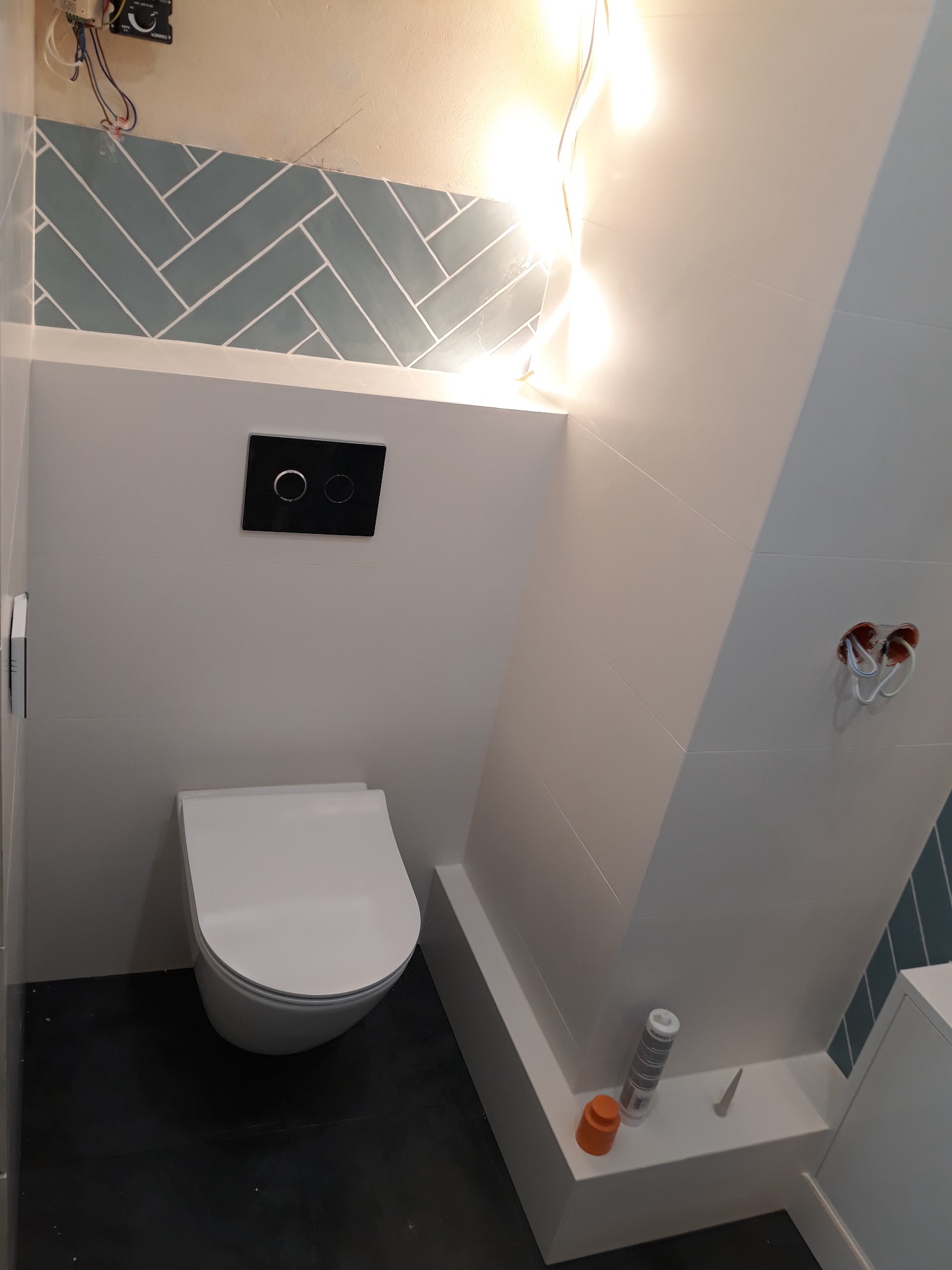 Nowoczesna toaleta wisząca w łazience z szarą podłogą, białymi płytkami i dekoracyjnym pasem płytek ułożonych w jodełkę, widoczne gniazdko elektryczne i silikon sanitarny.