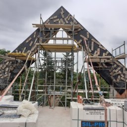 Przedsiębiorstwo Budowlane WB S.C Michał Wronkowski Łukasz Boczoń - Konstrukcja szczytu dachu w trakcie budowy, widoczne rusztowania, czarna membrana z przytwierdzonymi listwami, białe bloczki silikatowe na pierwszym planie.