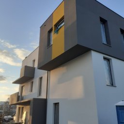 Przedsiębiorstwo Budowlane WB S.C Michał Wronkowski Łukasz Boczoń - Wielopiętrowy budynek w trakcie budowy z elementami wykończenia w kolorze białym, szarym i żółtym, okna zabezpieczone folią, widoczne materiały budowlane i przenośna toaleta.