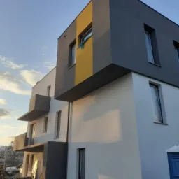 Wielopiętrowy budynek w trakcie budowy z elementami wykończenia w kolorze białym, szarym i ż&oacute;łtym, okna zabezpieczone folią, widoczne materiały budowlane i przenośna toaleta.