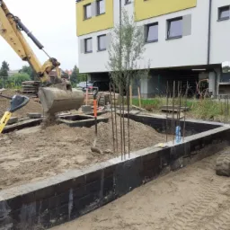 Wykop pod fundamenty z częściowo wylaną betonową konstrukcją, wzmocnioną stalowymi prętami, z żółtą koparką w trakcie zasypywania wykopu piaskiem, na tle budynku mieszkalnego.