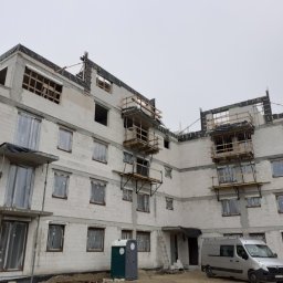 Przedsiębiorstwo Budowlane WB S.C Michał Wronkowski Łukasz Boczoń - Wielopiętrowy budynek w trakcie budowy z widocznymi rusztowaniami, niezabezpieczonymi oknami, przenośną toaletą i zaparkowanym vanem na placu budowy w pochmurny dzień.