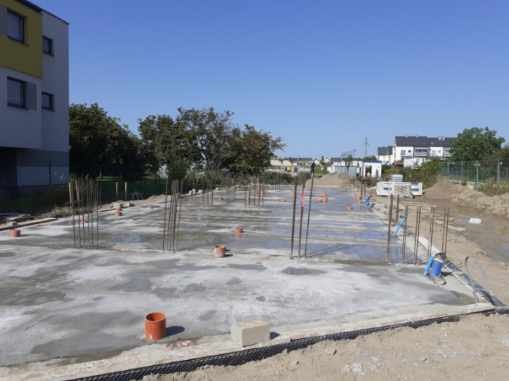 Wylany fundament z wystającymi prętami zbrojeniowymi, widoczne pomarańczowe rury i elementy szalunku, w tle budynek mieszkalny i zabudowa osiedlowa.