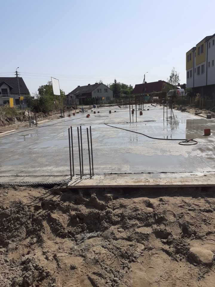 Wylany, świeży fundament z wystającymi prętami zbrojeniowymi, widoczne elementy instalacji i otaczający teren z ziemią.