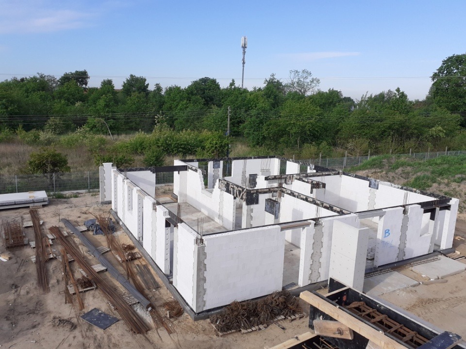 Wznoszenie ścian parterowego domu z białych bloczków, widoczne zbrojenia nad otworami okiennymi i drzwiami, na zewnątrz składowane pręty zbrojeniowe, w tle zieleń i maszt telekomunikacyjny.