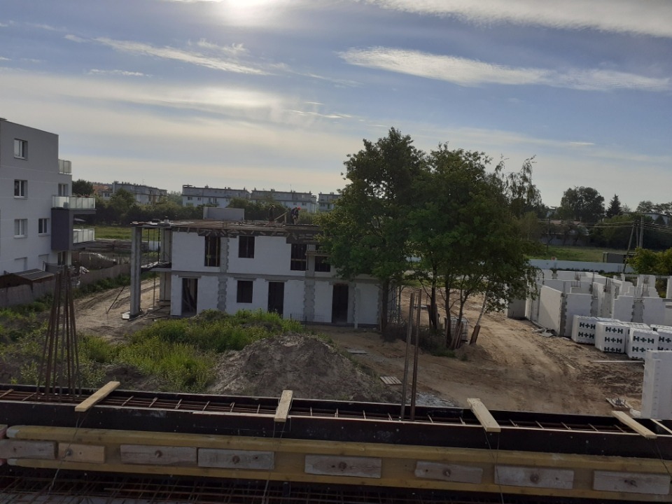 Wznoszona dwukondygnacyjna konstrukcja budynku z betonu komórkowego, widok z góry na szalunek stropu, w tle stosy bloczków H+H oraz zieleń i zabudowa osiedlowa pod błękitnym niebem.