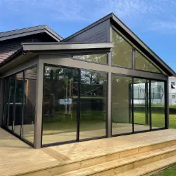 Nowoczesna przeszklona dobud&oacute;wka do domu z drewnianym tarasem i schodami, widok z zewnątrz na tle zielonego trawnika i błękitnego nieba.