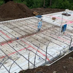 Przygotowanie płyty fundamentowej: izolacja termiczna, zbrojenie siatką stalową i rozprowadzone rury ogrzewania podłogowego, widoczne również pionowe rury instalacyjne i laserowe wyznaczenie poziomu.