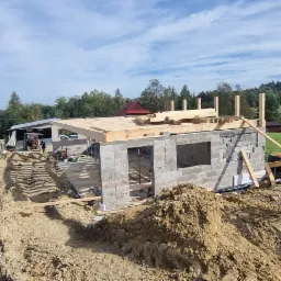 Budowa domu z bloczków betonowych w trakcie realizacji, widoczna konstrukcja dachu z drewnianych belek, w tle krajobraz wiejski i traktor.