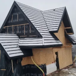 Budowa domu z widoczną konstrukcją dachu pokrytą czarną folią i częściowo dachówką, ośnieżoną. Ściany obłożone płytami drewnopochodnymi. W tle zabudowa mieszkalna i zalesione wzgórza w zimowej...