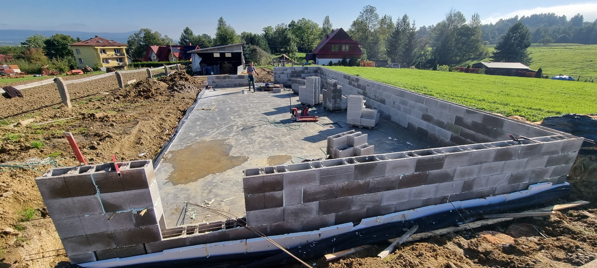 Wznoszenie ścian fundamentowych z bloczków betonowych na izolowanej płycie fundamentowej w otoczeniu krajobrazu wiejskiego.