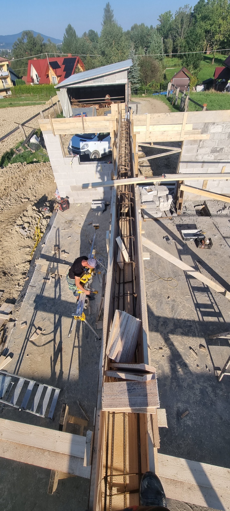 Pracownik na budowie, z góry, tnie drewno na kozłach przy szalunku zbrojonym stalą, w tle budynki mieszkalne i pojazd.