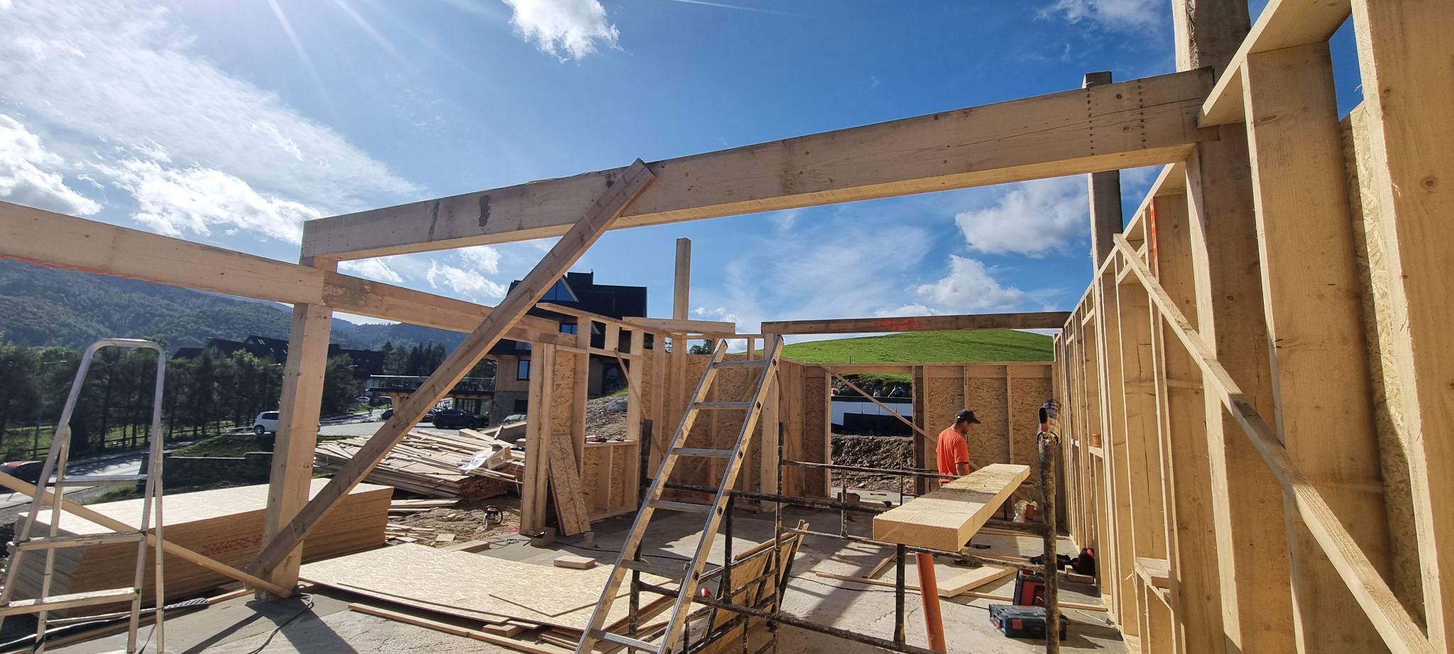 Wznoszenie drewnianej konstrukcji domu szkieletowego z widokiem na góry w tle, widoczna drabina i pracownik w pomarańczowej koszulce.