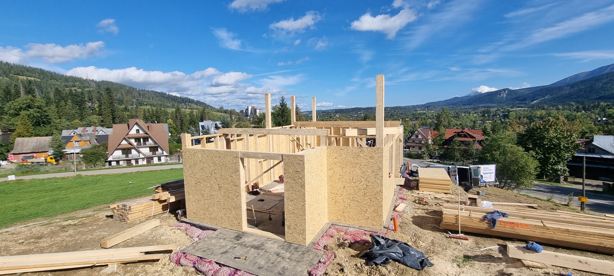 Konstrukcja drewniana domu w trakcie budowy, widok na drewniane ściany z płyt OSB, w tle góry i zabudowa mieszkalna.
