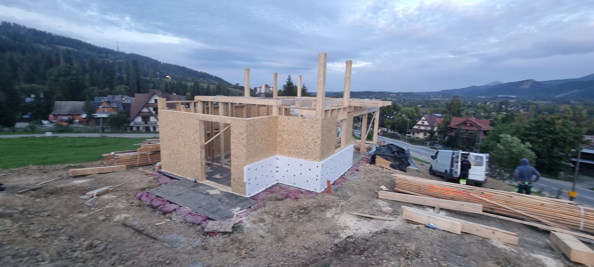 Wznoszenie konstrukcji domu szkieletowego z płyt OSB, widoczne izolacja fundamentów oraz górzysty krajobraz w tle, częściowo pochmurne niebo.