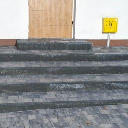 BRUKART - Schody wejściowe z szarej kostki brukowej prowadzące do drzwi garażowych, obok żółta skrzynka gazowa.