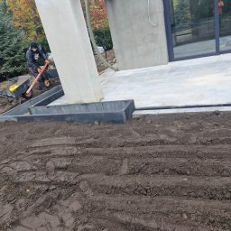 BRUKART - Pracownik z taczką i poziomicą przygotowuje obrzeże tarasu z szarych betonowych krawężników na ubitej ziemi przed nowoczesnym domem z dużymi oknami.